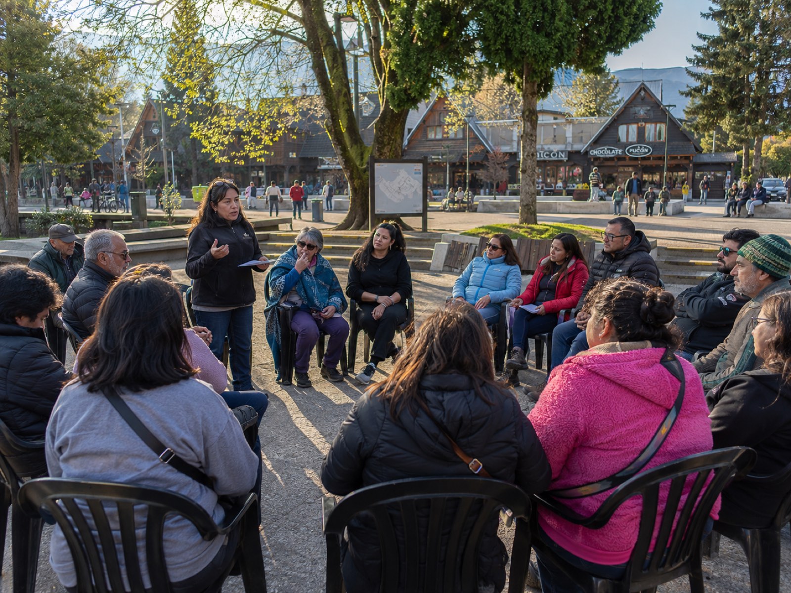 Una funcionaria municipal expone de pie ante un círculo de vecinos sentados en la plaza de Pucón, en una asamblea al aire libre